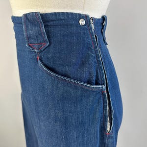 May include: High-waisted dark blue denim pants featuring a side zipper closure, a single silver-toned metal button at the waist, and prominent red contrast stitching along the pocket edges and seams, displayed on a mannequin torso.