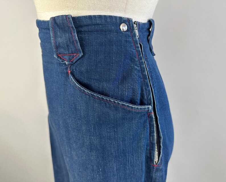 May include: High-waisted dark blue denim pants featuring a side zipper closure, a single silver-toned metal button at the waist, and prominent red contrast stitching along the pocket edges and seams, displayed on a mannequin torso.