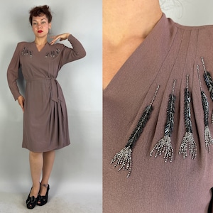 May include: A mauve-colored dress with long sleeves and a wrap-style waist. The dress features decorative beaded accents near the neckline. The dress is knee-length and is paired with black heels.