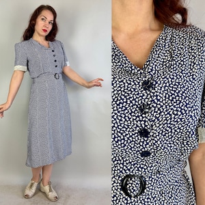 May include: A blue and white floral print dress with a white lace trim on the sleeves and a black belt. The dress has a button-up front and a knee-length skirt.