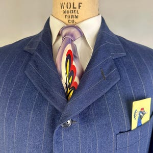 Puede incluir: Una americana azul de rayas diplom&aacute;ticas con una camisa blanca de cuello y una corbata con un dise&ntilde;o abstracto en morado, rojo y amarillo. Un pa&ntilde;uelo de bolsillo amarillo con el dibujo de un pavo real con sombrero rojo en el bolsillo.