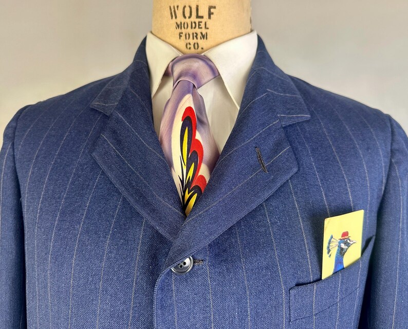 Puede incluir: Una americana azul de rayas diplom&aacute;ticas con una camisa blanca de cuello y una corbata con un dise&ntilde;o abstracto en morado, rojo y amarillo. Un pa&ntilde;uelo de bolsillo amarillo con el dibujo de un pavo real con sombrero rojo en el bolsillo.