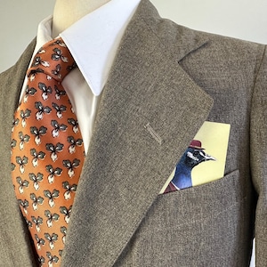 Op de afbeelding: Een bruine blazer met textuur gedragen over een wit overhemd met kraag en een oranje das met een herhalend donkergroen en wit bloemenpatroon. Een pochet met een grafische illustratie van een vogel met een rode hoed zit in de borstzak.