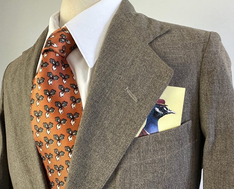 Op de afbeelding: Een bruine blazer met textuur gedragen over een wit overhemd met kraag en een oranje das met een herhalend donkergroen en wit bloemenpatroon. Een pochet met een grafische illustratie van een vogel met een rode hoed zit in de borstzak.