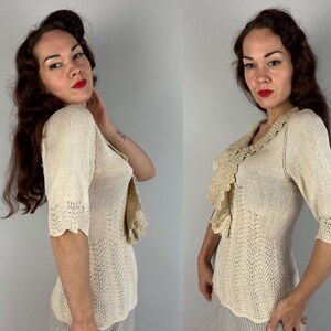 May include: A cream-colored knitted sweater with a ruffled neckline and short sleeves. The sweater is made of a delicate, open-weave fabric.