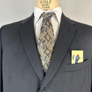 May include: A dark charcoal blazer worn over a crisp white collared shirt with a patterned necktie featuring geometric shapes in shades of tan, green, and black. A small yellow pocket square with a peacock illustration is tucked in the pocket.