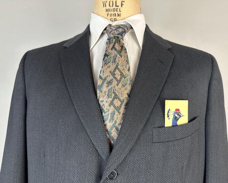 May include: A dark charcoal blazer worn over a crisp white collared shirt with a patterned necktie featuring geometric shapes in shades of tan, green, and black. A small yellow pocket square with a peacock illustration is tucked in the pocket.
