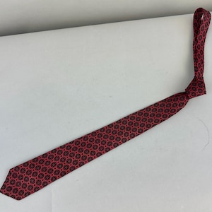 May include: A red necktie with a repeating black floral pattern. The tie is knotted and lies diagonally across a white surface. The tie's design features small, circular black floral motifs.