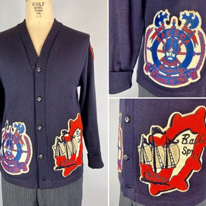 May include: Navy blue vintage cardigan sweater with a V-neck and button closure. The sweater features embroidered patches with nautical and school-themed designs in red, white, and blue. Patches include text that reads "Balboa" and "Class of 1961".