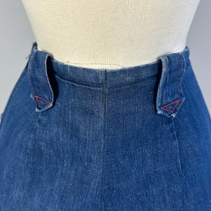 May include: High-waisted dark blue denim pants featuring a structured waistband with two prominent belt loops. Each belt loop is reinforced with distinct red stitching at the base, creating a decorative triangular pattern against the deep indigo fabric.