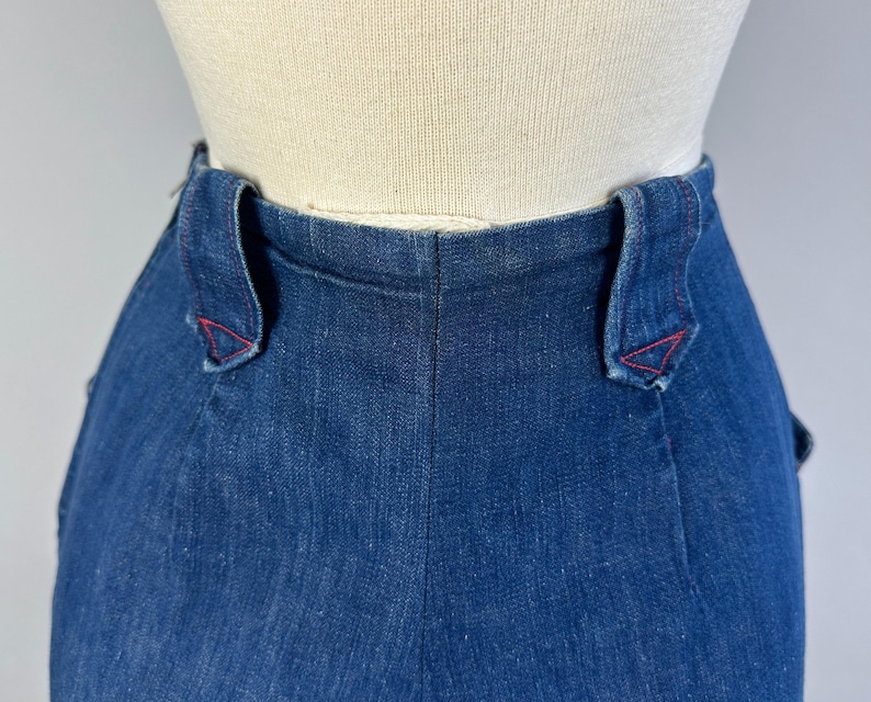 May include: High-waisted dark blue denim pants featuring a structured waistband with two prominent belt loops. Each belt loop is reinforced with distinct red stitching at the base, creating a decorative triangular pattern against the deep indigo fabric.