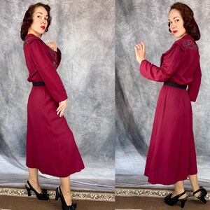 May include: A burgundy dress with long sleeves, a black belt, and a midi-length skirt. The dress has embroidered detailing on the shoulders and collar. The model is wearing black heels with a bow detail. The background is a grey and white backdrop.