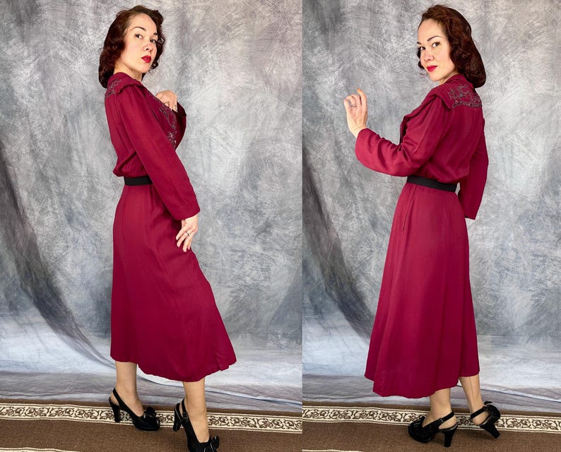 May include: A burgundy dress with long sleeves, a black belt, and a midi-length skirt. The dress has embroidered detailing on the shoulders and collar. The model is wearing black heels with a bow detail. The background is a grey and white backdrop.