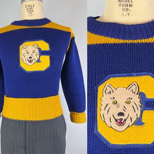 May include: A blue and gold knitted sweater with a wolf's head and letter C embroidered on the front. The sweater has a ribbed collar, cuffs, and hem.