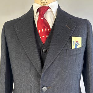 May include: A charcoal gray suit jacket with a subtle pinstripe pattern, worn over a white collared shirt and a dark vest. A red tie with a white geometric pattern is tied at the neck. A yellow pocket square features a blue peacock illustration.