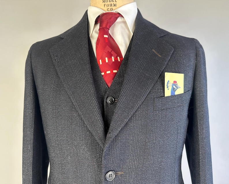 May include: A charcoal gray suit jacket with a subtle pinstripe pattern, worn over a white collared shirt and a dark vest. A red tie with a white geometric pattern is tied at the neck. A yellow pocket square features a blue peacock illustration.