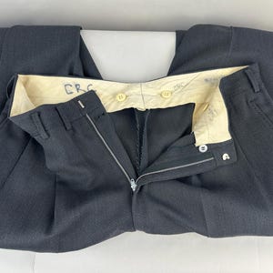 May include: Dark charcoal gray trousers featuring a textured fabric, a metal zipper fly, and a hook-and-bar closure. The interior waistband is lined with cream-colored fabric, displaying the handwritten letters CRC in blue ink and two light-colored buttons.