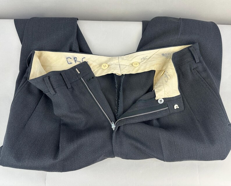 May include: Dark charcoal gray trousers featuring a textured fabric, a metal zipper fly, and a hook-and-bar closure. The interior waistband is lined with cream-colored fabric, displaying the handwritten letters CRC in blue ink and two light-colored buttons.