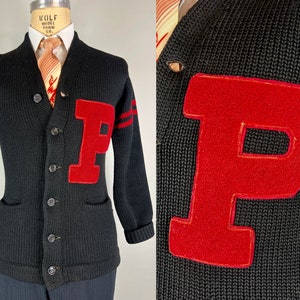 Puede incluir: Cárdigan negro de punto con un gran parche rojo con la letra "P" en el pecho. El suéter tiene cuello chal, cierre de botones y dos bolsillos delanteros. Rayas rojas adornan la manga izquierda. Estilo vintage.