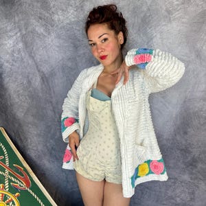 May include: A white textured cardigan featuring long sleeves with blue and pink floral accents at the cuffs and hem. The garment has a front pocket and is worn over a light-colored polka dot romper with a blue bodice. The fabric has a raised, bumpy surface.