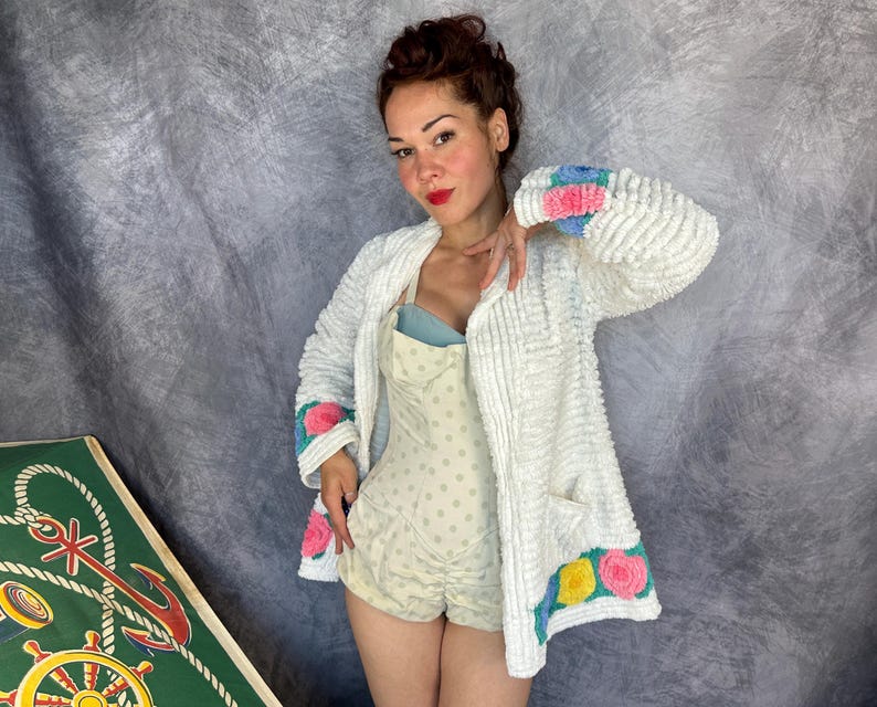 May include: A white textured cardigan featuring long sleeves with blue and pink floral accents at the cuffs and hem. The garment has a front pocket and is worn over a light-colored polka dot romper with a blue bodice. The fabric has a raised, bumpy surface.