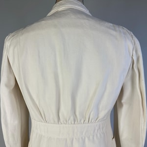 May include: A cream-coloured jacket featuring a structured collar, long sleeves, and a fitted waistline with subtle vertical pleating detail. The garment is crafted from a solid, light-toned fabric and displays a clean, tailored construction throughout.