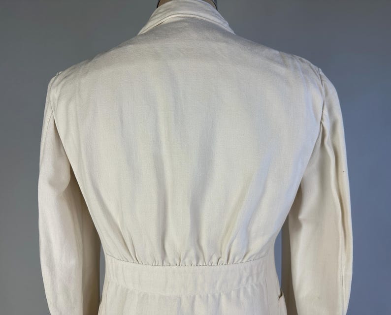 May include: A cream-coloured jacket featuring a structured collar, long sleeves, and a fitted waistline with subtle vertical pleating detail. The garment is crafted from a solid, light-toned fabric and displays a clean, tailored construction throughout.