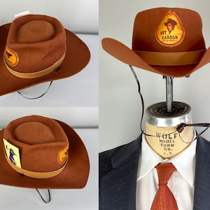 May include: A brown felt cowboy hat with a brown leather band and a patch featuring a drawing of Kit Carson. The hat is on a mannequin head with a brown leather chin strap.