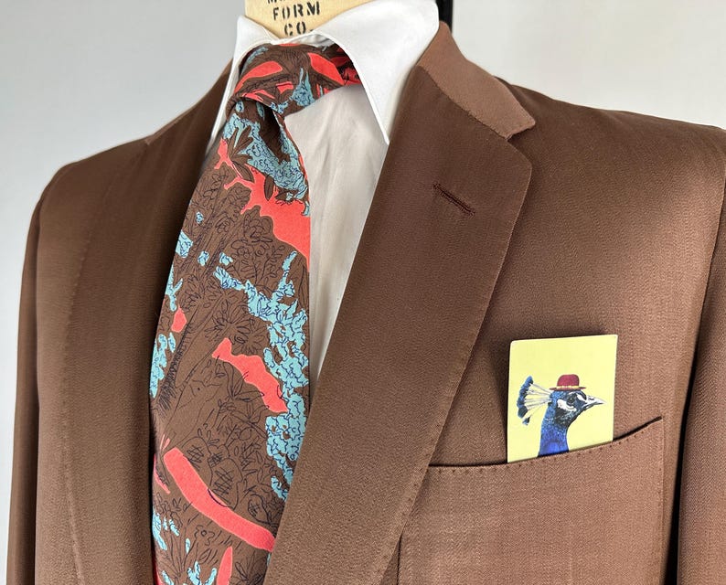 May include: A brown suit jacket with a white collared shirt and a patterned tie. The tie features a design with brown, red, and blue elements. A small card with a peacock illustration in a pocket adds a pop of color.