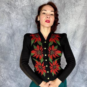 May include: Black cardigan sweater with a V-neck and long sleeves. The sweater features a poinsettia floral design in red and green, with gold-colored buttons. The sleeves have a brown stripe detail.