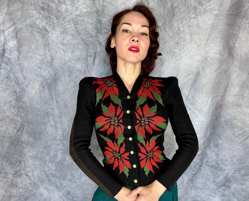May include: Black cardigan sweater with a V-neck and long sleeves. The sweater features a poinsettia floral design in red and green, with gold-colored buttons. The sleeves have a brown stripe detail.