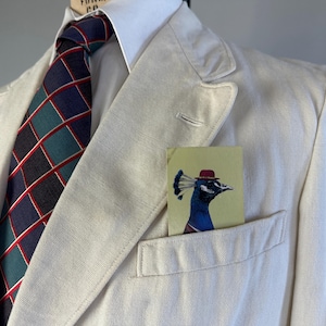 May include: A cream-coloured blazer worn with a white collared shirt and a dark blue tie featuring a teal and red grid pattern. A rectangular pocket square with a graphic illustration of a peacock wearing a small red hat sits in the chest pocket.