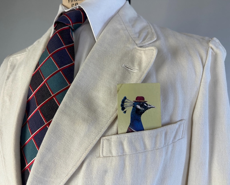 May include: A cream-coloured blazer worn with a white collared shirt and a dark blue tie featuring a teal and red grid pattern. A rectangular pocket square with a graphic illustration of a peacock wearing a small red hat sits in the chest pocket.