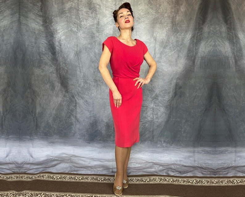 May include: A vibrant red knee-length dress featuring short cap sleeves, a rounded neckline, and a fitted bodice with subtle draping at the waist. The skirt follows a straight silhouette, ending just below the knee, creating a clean and structured look.