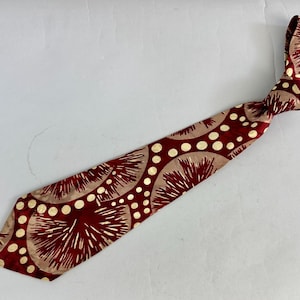 May include: A maroon tie featuring a repeating pattern of beige circles and white radial designs. The tie is knotted at the top and lies diagonally across a white surface. The tie's design combines geometric and floral elements.