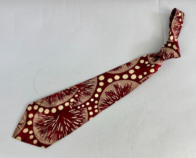 May include: A maroon tie featuring a repeating pattern of beige circles and white radial designs. The tie is knotted at the top and lies diagonally across a white surface. The tie's design combines geometric and floral elements.
