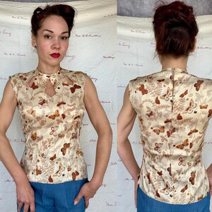 May include: A sleeveless, cream-colored top with a butterfly and foliage pattern. The top features a keyhole neckline and a high collar. The butterflies are in shades of brown and orange. The top is made of a silky material.