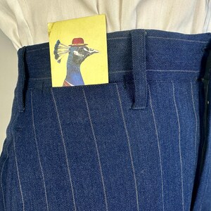 Puede incluir: Pantalones azul oscuro de talle alto con rayas diplom&aacute;ticas blancas finas y trabillas para cintur&oacute;n. Una tarjeta rectangular amarilla con la ilustraci&oacute;n de un pavo real con un bomb&iacute;n rojo asoma por el bolsillo delantero sobre una camisa blanca.