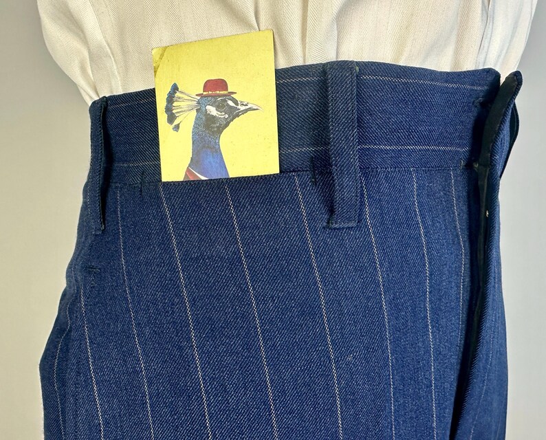 Puede incluir: Pantalones azul oscuro de talle alto con rayas diplom&aacute;ticas blancas finas y trabillas para cintur&oacute;n. Una tarjeta rectangular amarilla con la ilustraci&oacute;n de un pavo real con un bomb&iacute;n rojo asoma por el bolsillo delantero sobre una camisa blanca.