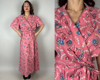 May include: A vintage pink wrap dress with a floral pattern. The dress features short sleeves, a ruffled collar, and a matching tie belt. The floral design includes blue and white accents.