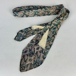 May include: A long, narrow necktie featuring a textured, multi-colored geometric pattern in shades of muted green, brown, and tan. The tie has a pointed end and a visible cream-colored lining on the interior, with loose threads near the seam.