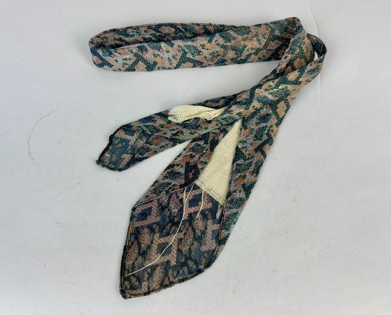 May include: A long, narrow necktie featuring a textured, multi-colored geometric pattern in shades of muted green, brown, and tan. The tie has a pointed end and a visible cream-colored lining on the interior, with loose threads near the seam.
