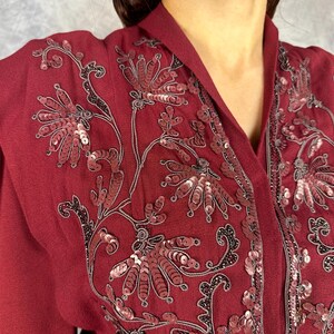 May include: A burgundy top with floral sequin embroidery. The design incorporates red and silver sequins with black detailing. The top has a V-neck and long sleeves. The fabric looks like a soft, flowing material.