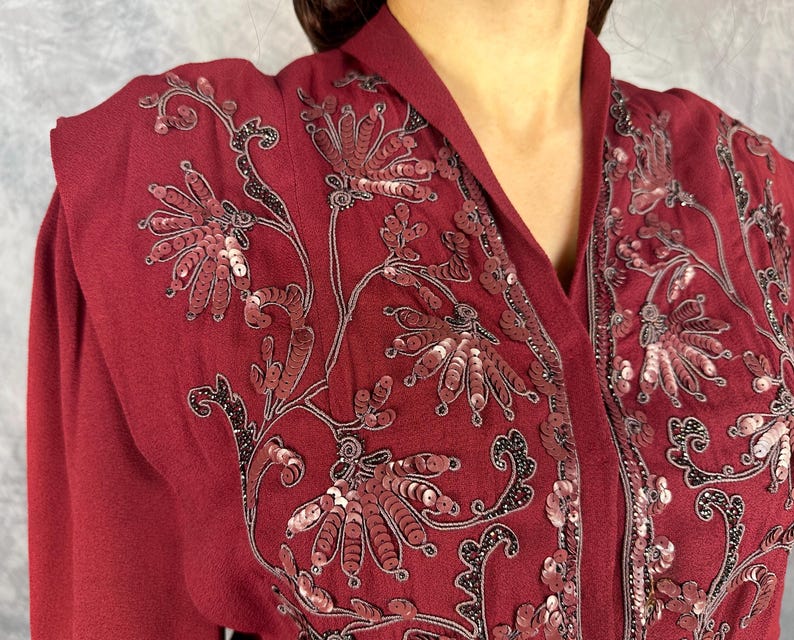 May include: A burgundy top with floral sequin embroidery. The design incorporates red and silver sequins with black detailing. The top has a V-neck and long sleeves. The fabric looks like a soft, flowing material.