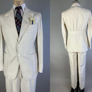 May include: A cream-coloured suit consisting of a two-button blazer with patch pockets and a breast pocket containing a small card, paired with matching trousers. The blazer features a cinched waist detail on the back and is worn with a white shirt.