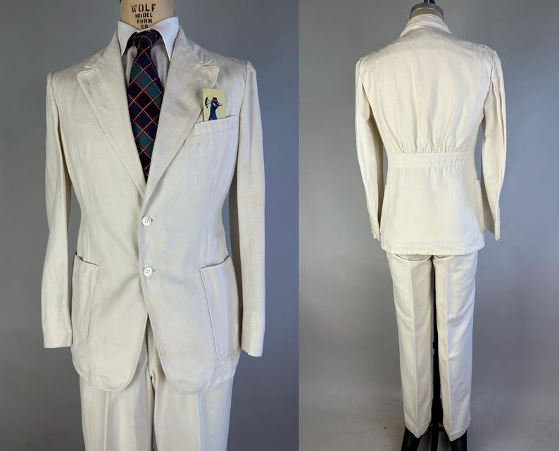 May include: A cream-coloured suit consisting of a two-button blazer with patch pockets and a breast pocket containing a small card, paired with matching trousers. The blazer features a cinched waist detail on the back and is worn with a white shirt.