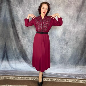 May include: A burgundy midi dress with long sleeves and a black belt. The bodice has detailed embroidery. The model is wearing black heels. The dress is a classic style.
