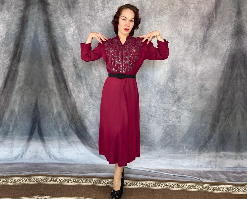 May include: A burgundy midi dress with long sleeves and a black belt. The bodice has detailed embroidery. The model is wearing black heels. The dress is a classic style.