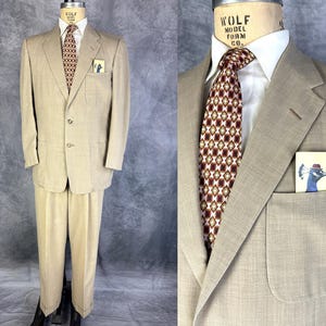 May include: A beige two-piece suit with a patterned tie and pocket square. The suit includes a two-button jacket and matching trousers. The tie has a geometric pattern in red, brown, and cream. A small card with a peacock illustration is in the breast pocket.