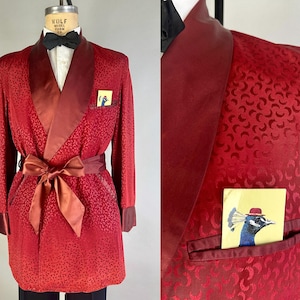 Könnte beinhalten: Eine dunkelrote Smokingjacke mit einem tonalen Halbmondmuster, einem Schalkragen aus Satin, passenden Satinmanschetten und einem Bindegürtel aus Satin. Ein kleines Einstecktuch mit einer Grafik eines Pfaus mit rotem Hut steckt in der Brusttasche.